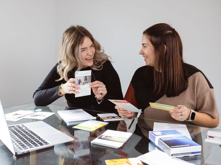Abbildung zwei Frauen unterhalten sich lachend über Karten die vor ihnen auf einem Tisch liegen
